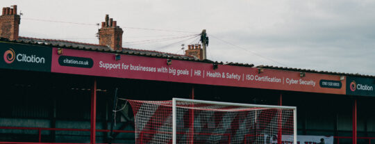 Picture of the Golf Road Stand at Altrincham FC with Citation's new branding. Stand image says 'Supporting businesses with big goals'